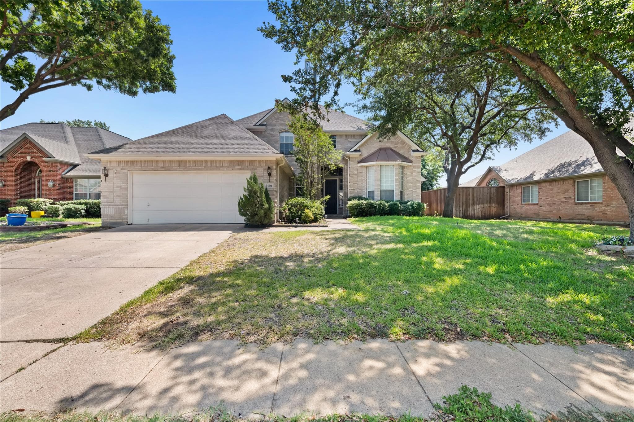 Front view of a two-story brick house with a garage, green lawn, and shady trees.