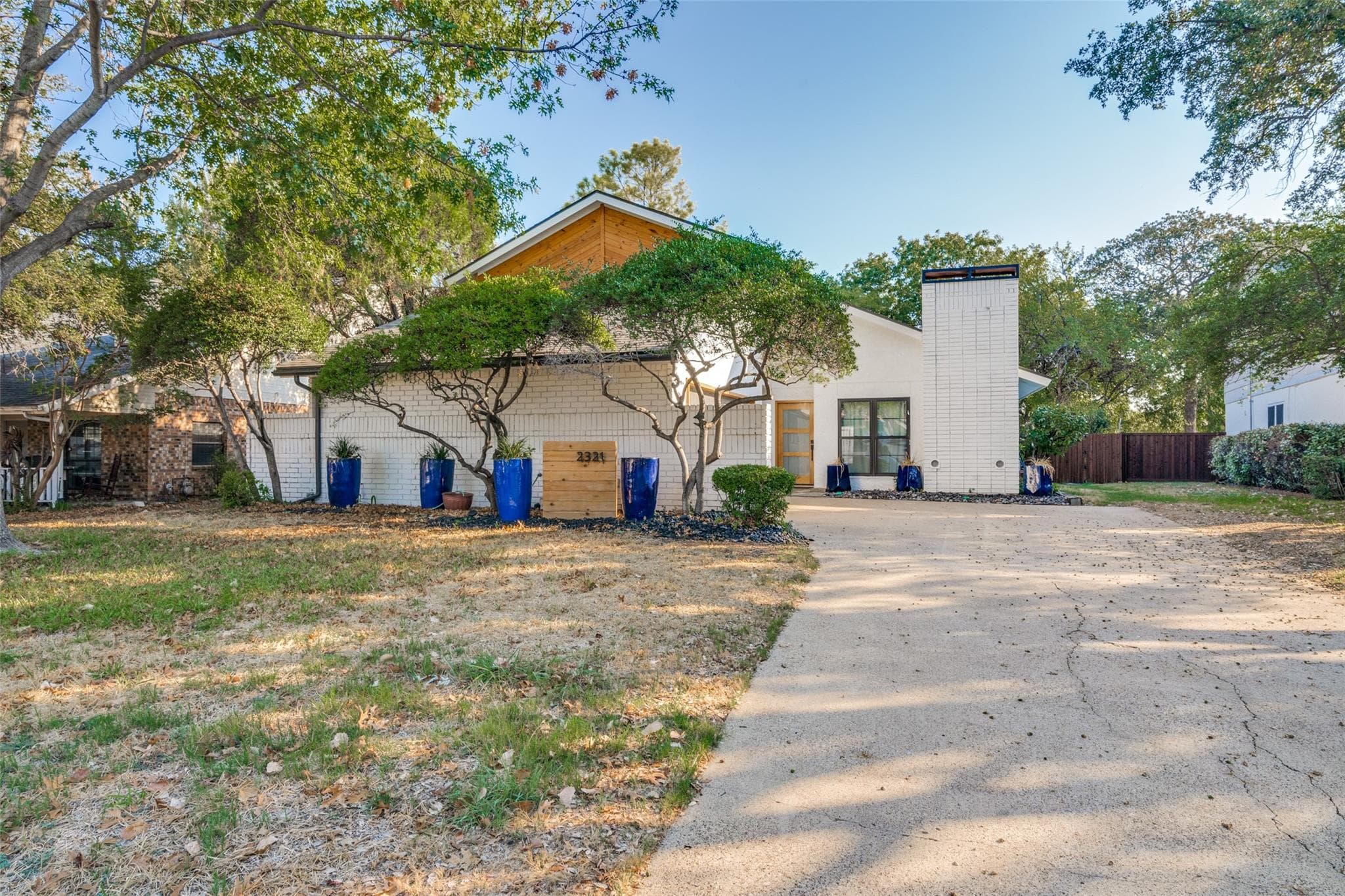 White brick house with wooden roof, blue planters, and a concrete driveway on a sunny day.